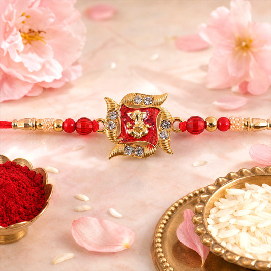 A red and gold Ganesha motif Rakhi with clear stones and red beads, surrounded by pink flowers and petals.