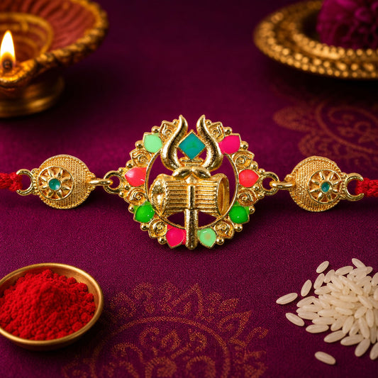 A gold Shiv Trishul Damru charm with pink, green, and blue stones on a red thread, surrounded by a lit diya and rice.