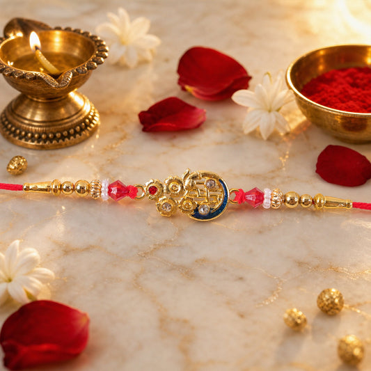 A golden finish designer Rakhi with red and white beads, a blue crescent, and a lit diya on a marble surface.
