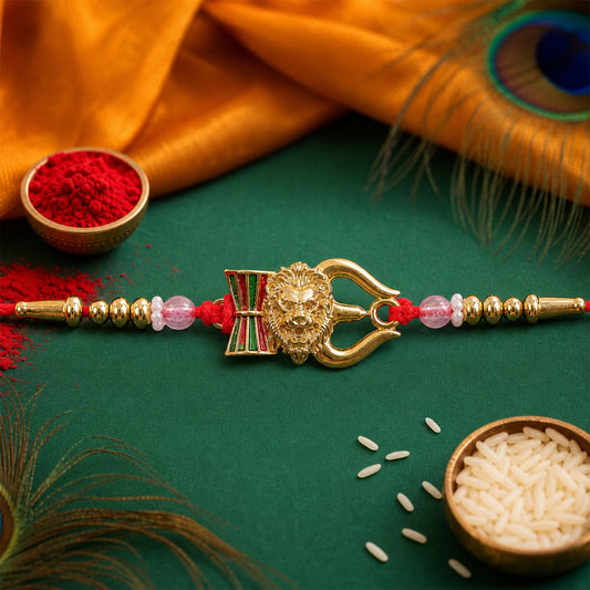 A red thread bracelet with a golden lion head, damaru, and trishul charm, flanked by gold and pink beads, on a green surface