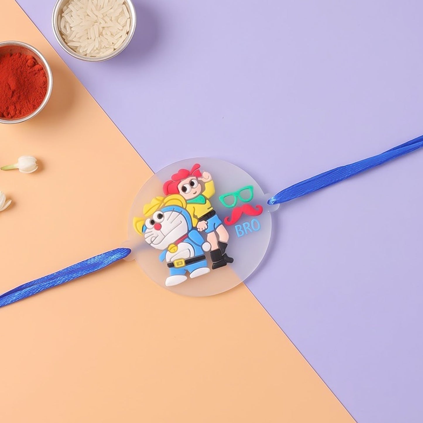 Blue Doraemon and Nobita Rakhi with a 'BRO' charm, on a split purple and orange background with puja thali items.