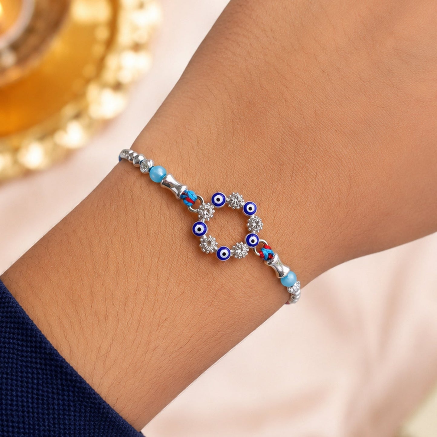 A blue and red thread rakhi with a circular evil eye charm, silver and turquoise beads on a person's wrist.