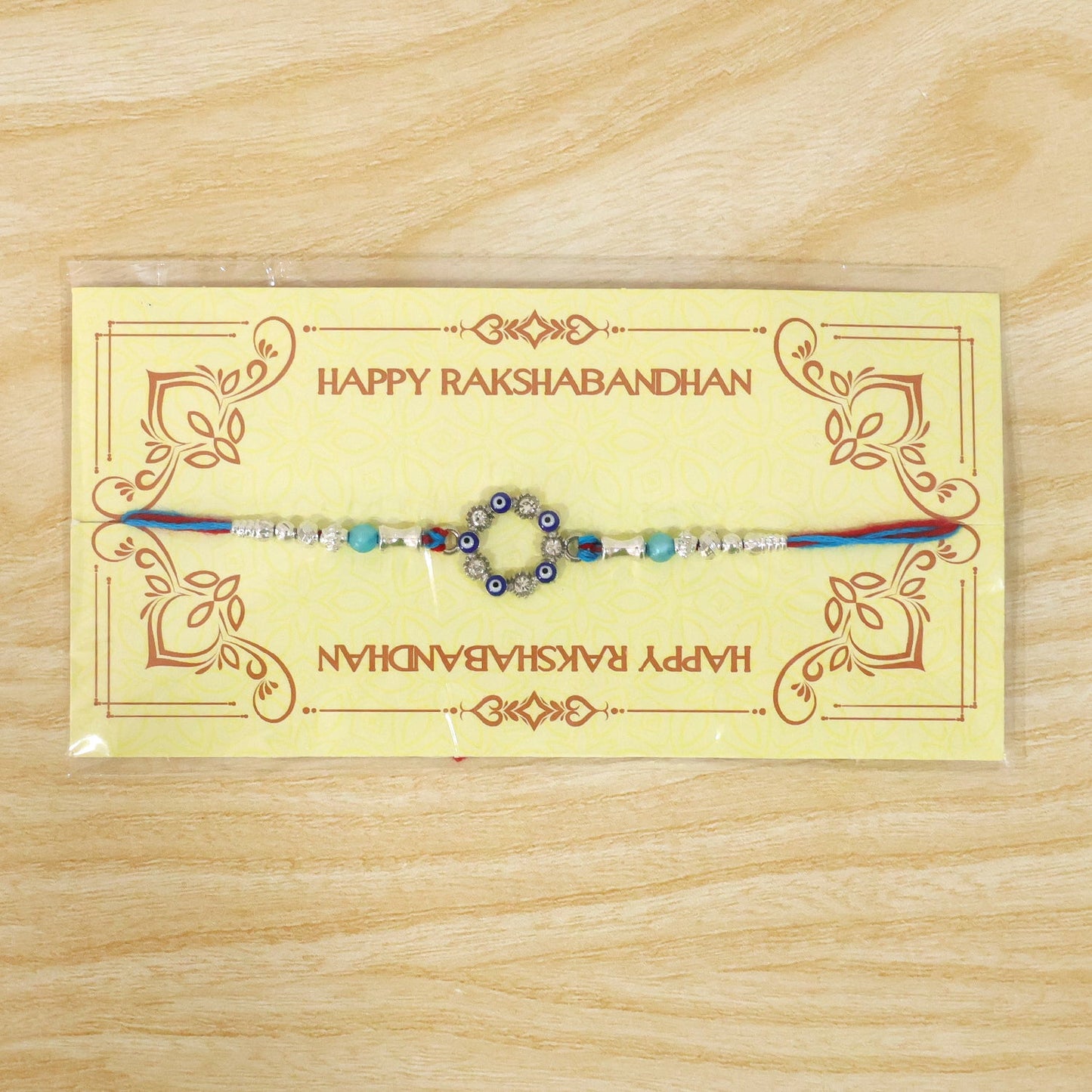 A blue and red thread rakhi with a circular evil eye charm, silver and turquoise beads on a yellow Happy Rakshabandhan card.