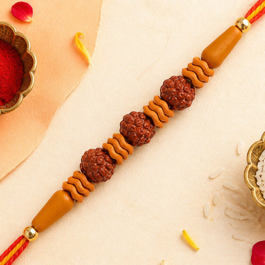 A Rudraksha Rakhi with three textured brown beads and wavy light brown spacers on a red and yellow thread, with a small bowl