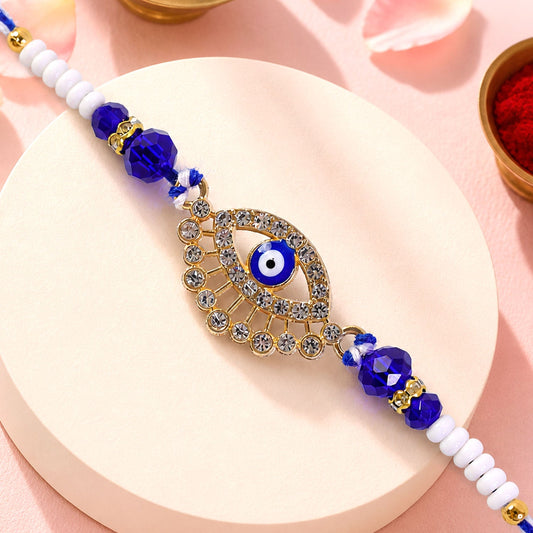A gold and diamond evil eye Rakhi with blue and white beads on a white surface with red powder.