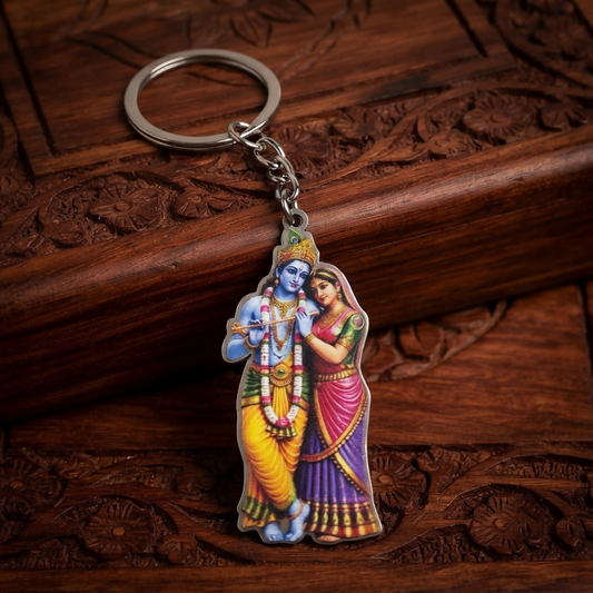 A vibrant Radha Krishna keychain with a silver ring rests on a dark carved wooden surface, showcasing the colorful divine