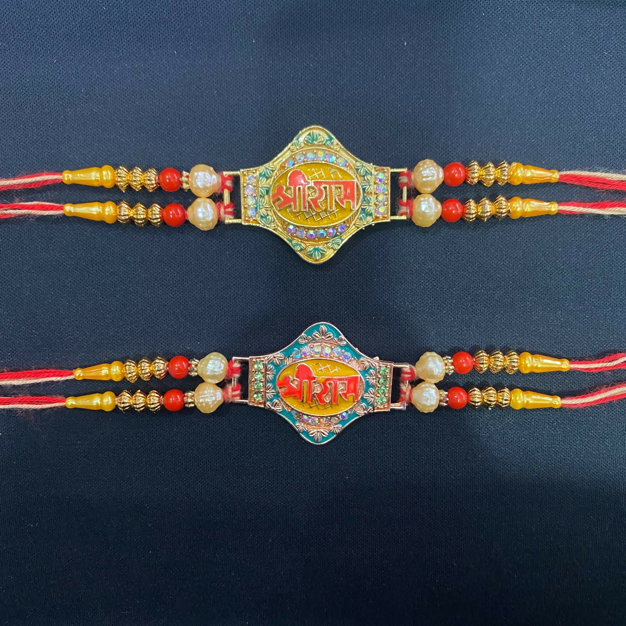 Two gold and pearl Rakhis with red and orange threads, one with a green center, the other blue, on a plain black background.