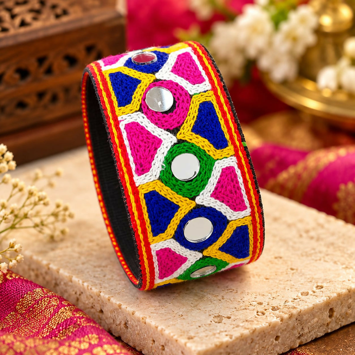 A vibrant cuff bracelet with mirror work and colorful embroidery sits on a light brown textured surface.