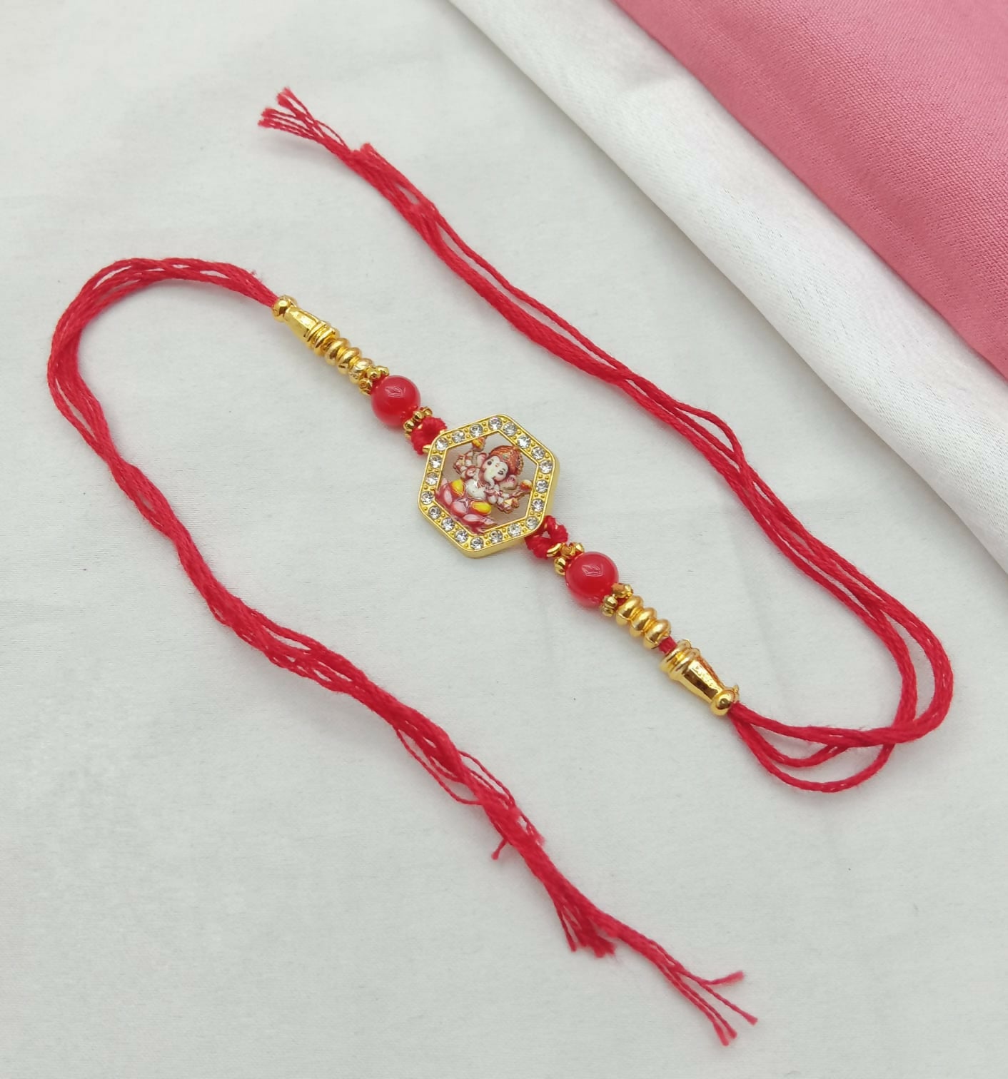 A red thread bracelet with a Ganesha figure charm and red beads lies on a white and pink fabric background.