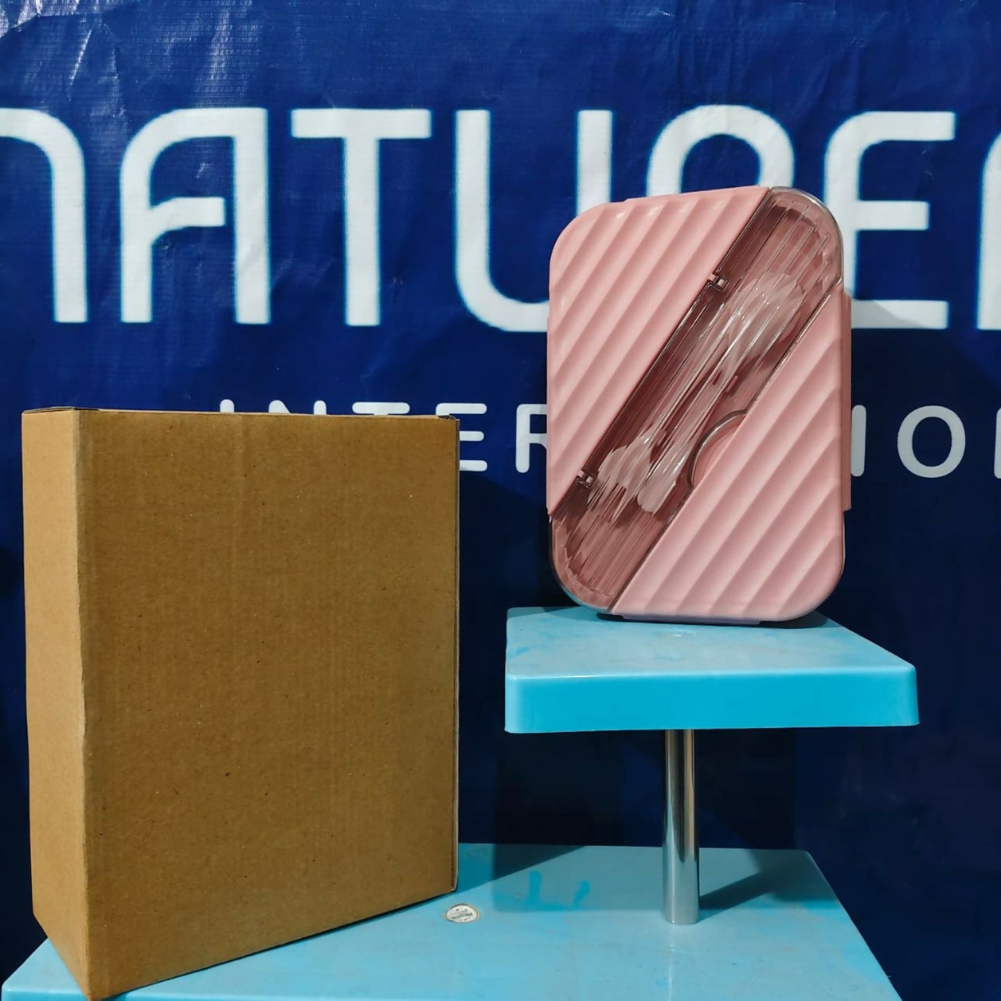 A pink bento lunch box with cutlery visible through its clear lid, standing upright next to a brown cardboard box.