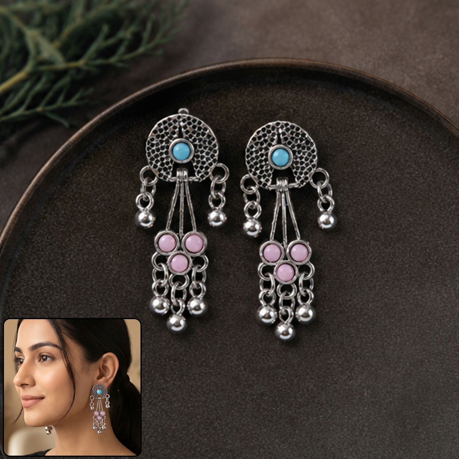 A pair of oxidised silver long drop earrings with pink and turquoise stones and ghungroo bells rest on a dark brown plate.