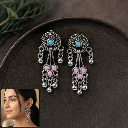 A pair of oxidised silver long drop earrings with pink and turquoise stones and ghungroo bells rest on a dark brown plate.