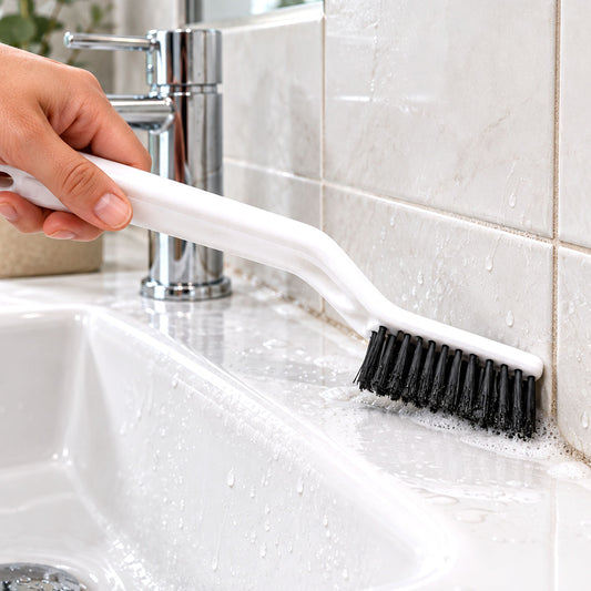 A hand holds a white brush with black bristles, cleaning a white tiled bathroom sink and wall.