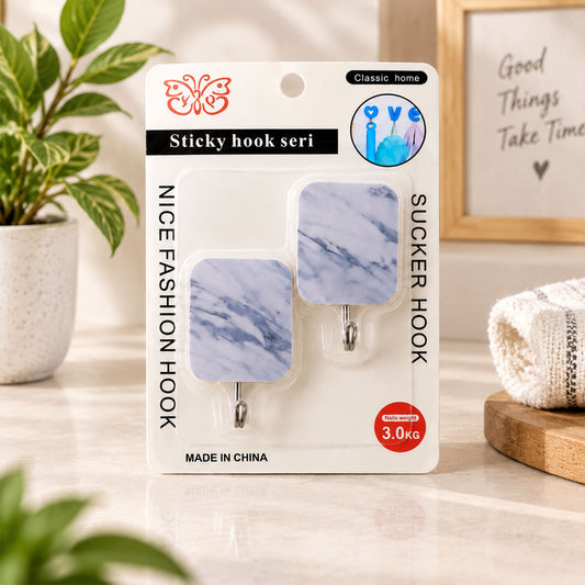 Two marble-patterned sticky hooks on retail packaging, displayed on a tiled counter next to a potted plant.