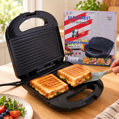 An open black sandwich maker with two grilled sandwiches inside, a spatula removing one, and its box in the background.