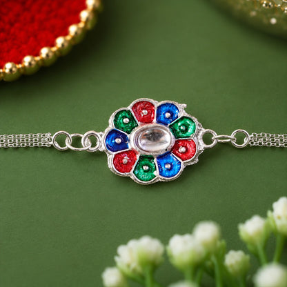 The silver chain bracelet with its vibrant enamel flower charm is displayed on a green surface, with soft white flowers in