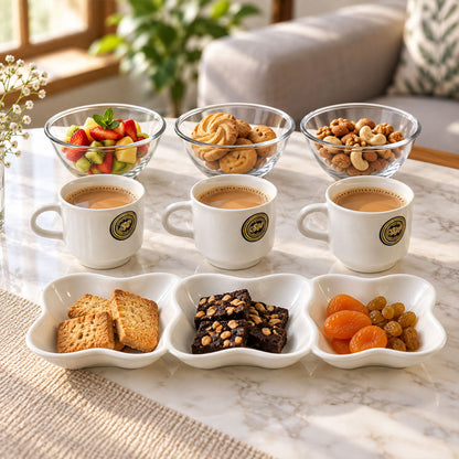 Three white ceramic mugs filled with a light brown beverage, three clear glass bowls containing fruit, cookies, and nuts,