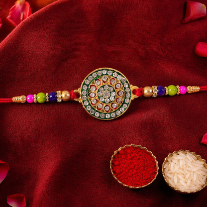 A gold and green circular Rakhi with clear stones and colorful beads lies on a red fabric with scattered rose petals and two
