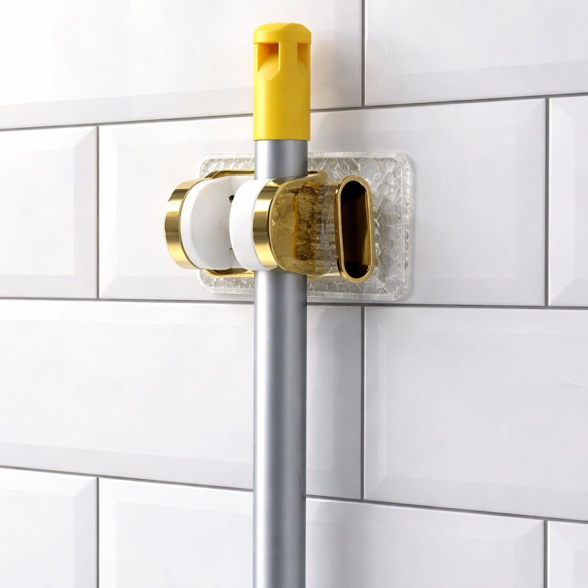 A gold and clear mop gripper holds a silver broom handle against a white tiled wall.