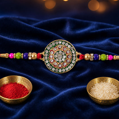 A gold and green enamel Rakhi with clear stones and colorful beads on a red thread, resting on a dark blue velvet surface.