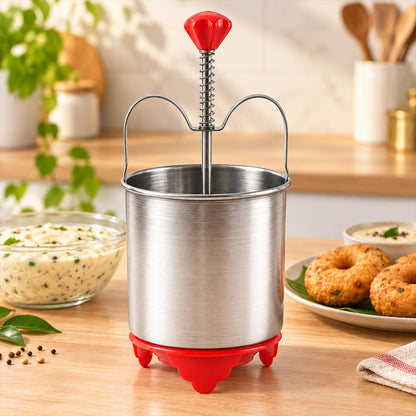 A stainless steel medu vada maker with a red handle and red base sits on a wooden counter next to a bowl of batter and a