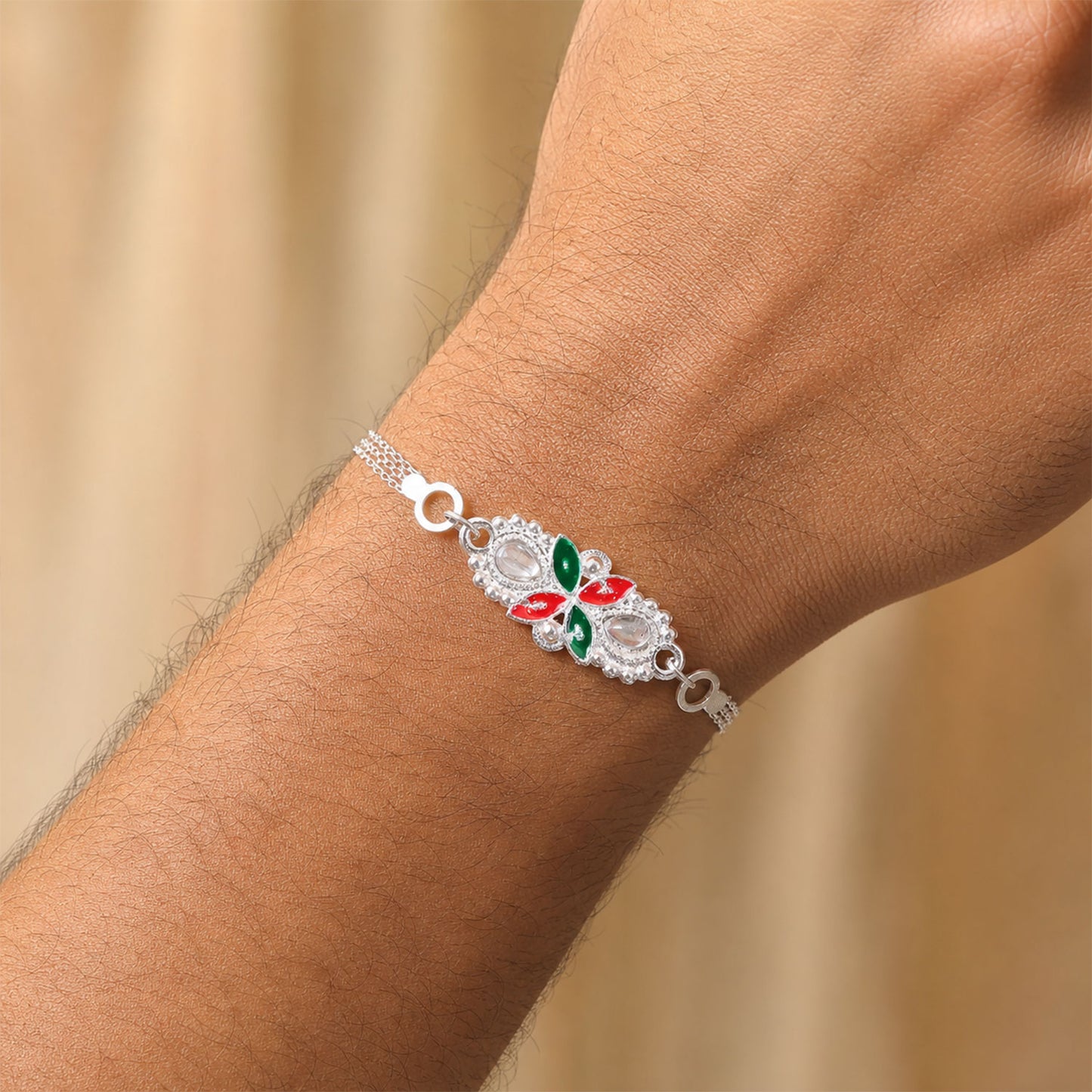 The silver-toned bracelet, adorned with red and green enamel and clear stones, is worn on a man's wrist, against a blurred