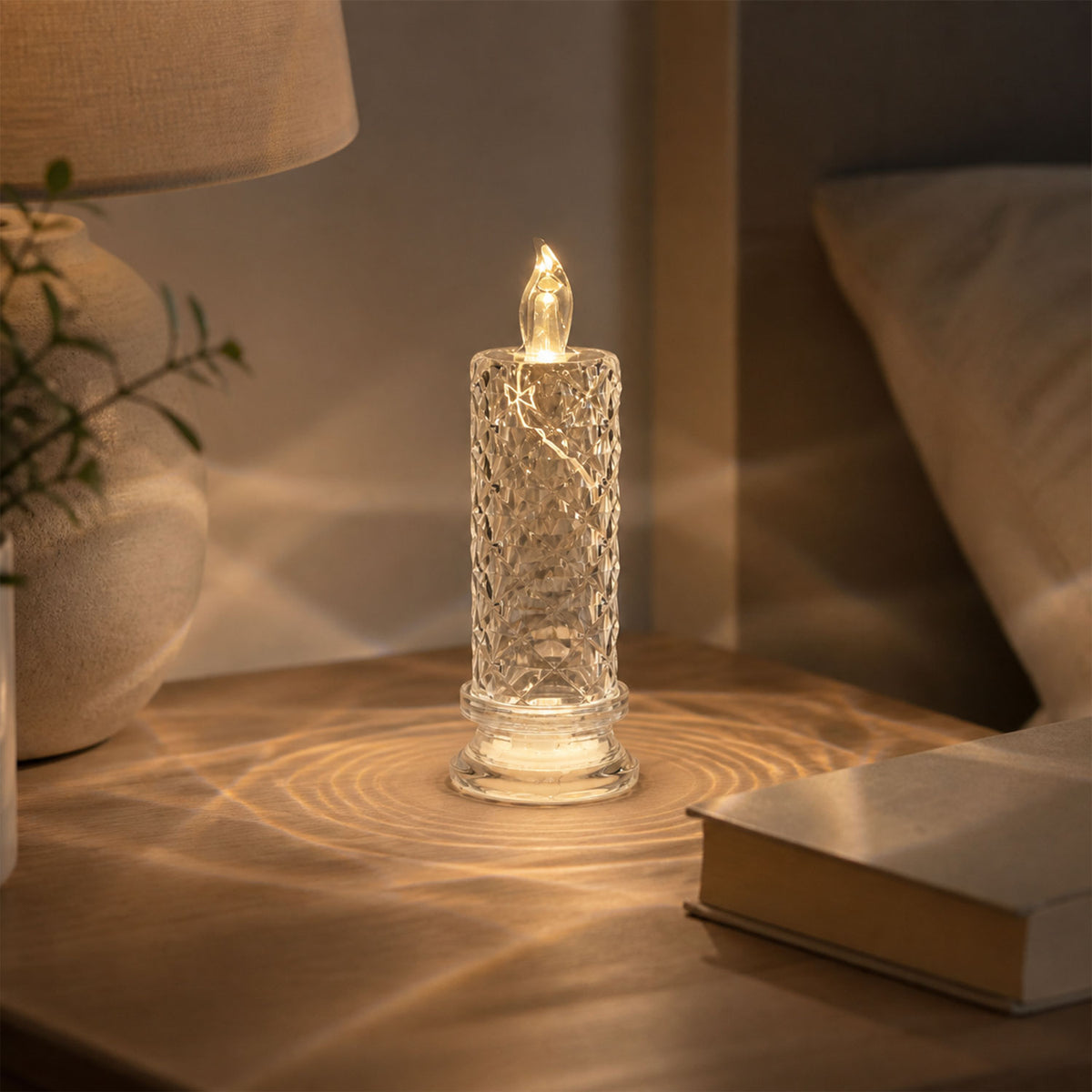 A clear, faceted crystal LED candle on a bedside table with a lamp and book, casting warm light.