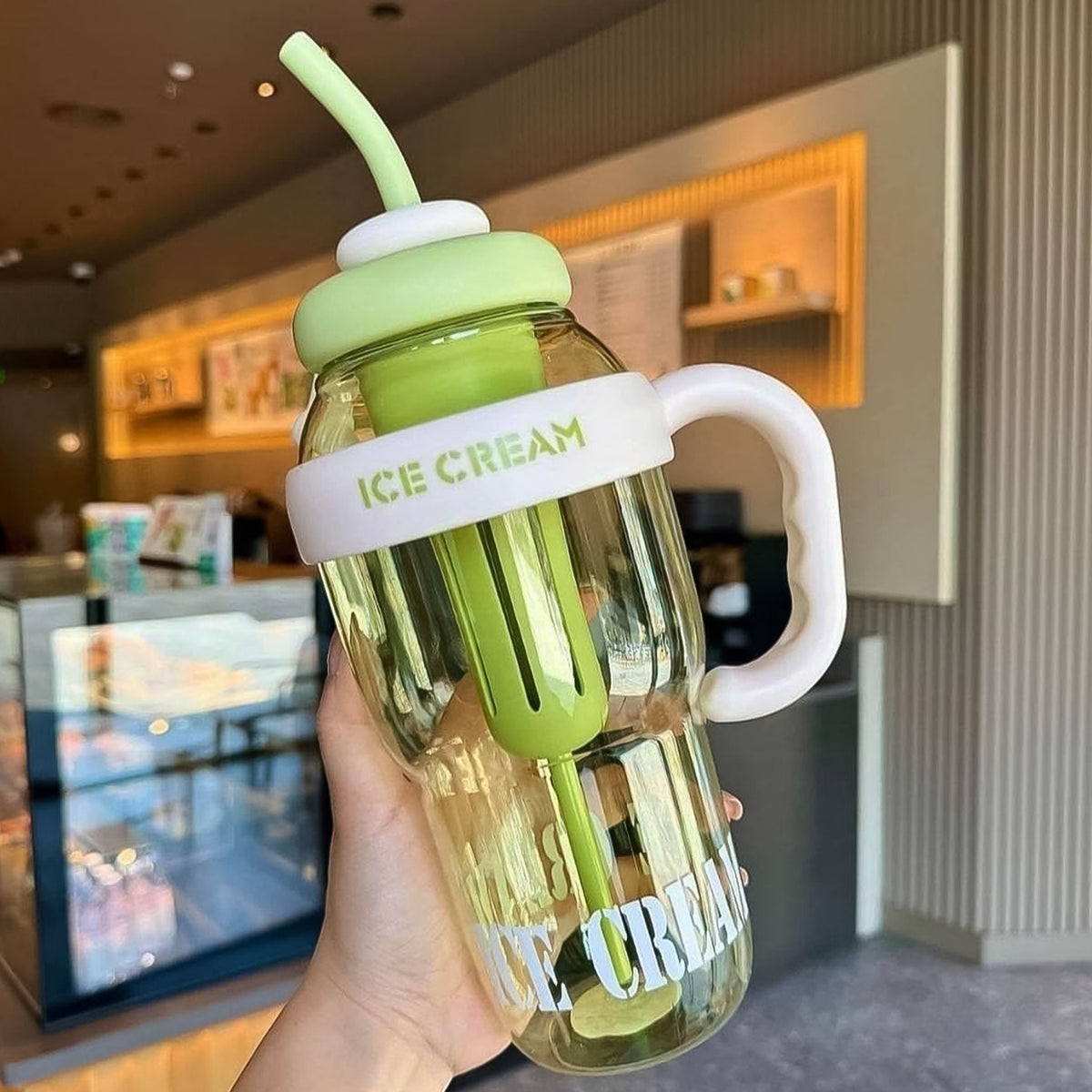 A hand holds a clear bottle with a green lid, straw, and handle, in a retail setting.