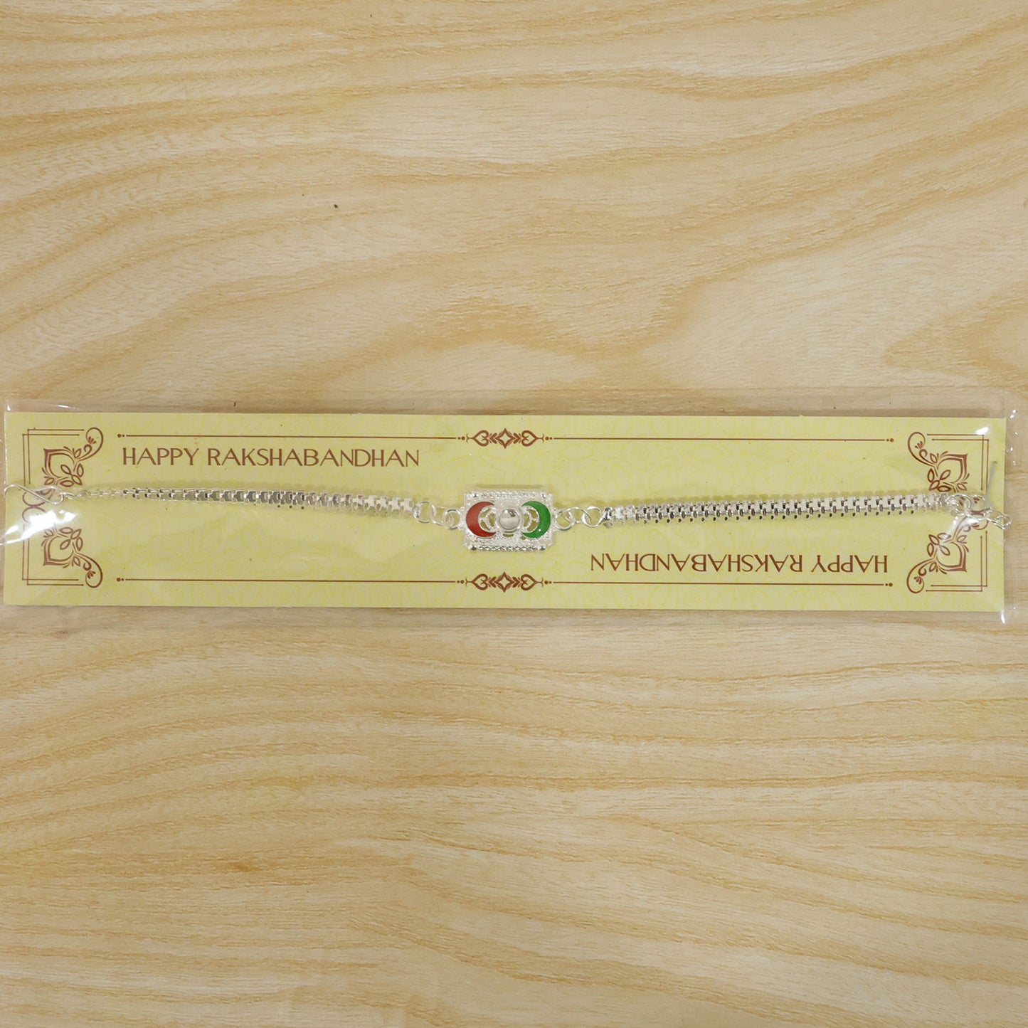 A silver-colored bracelet with a rectangular centerpiece featuring red and green crescent moon shapes, packaged on a yellow