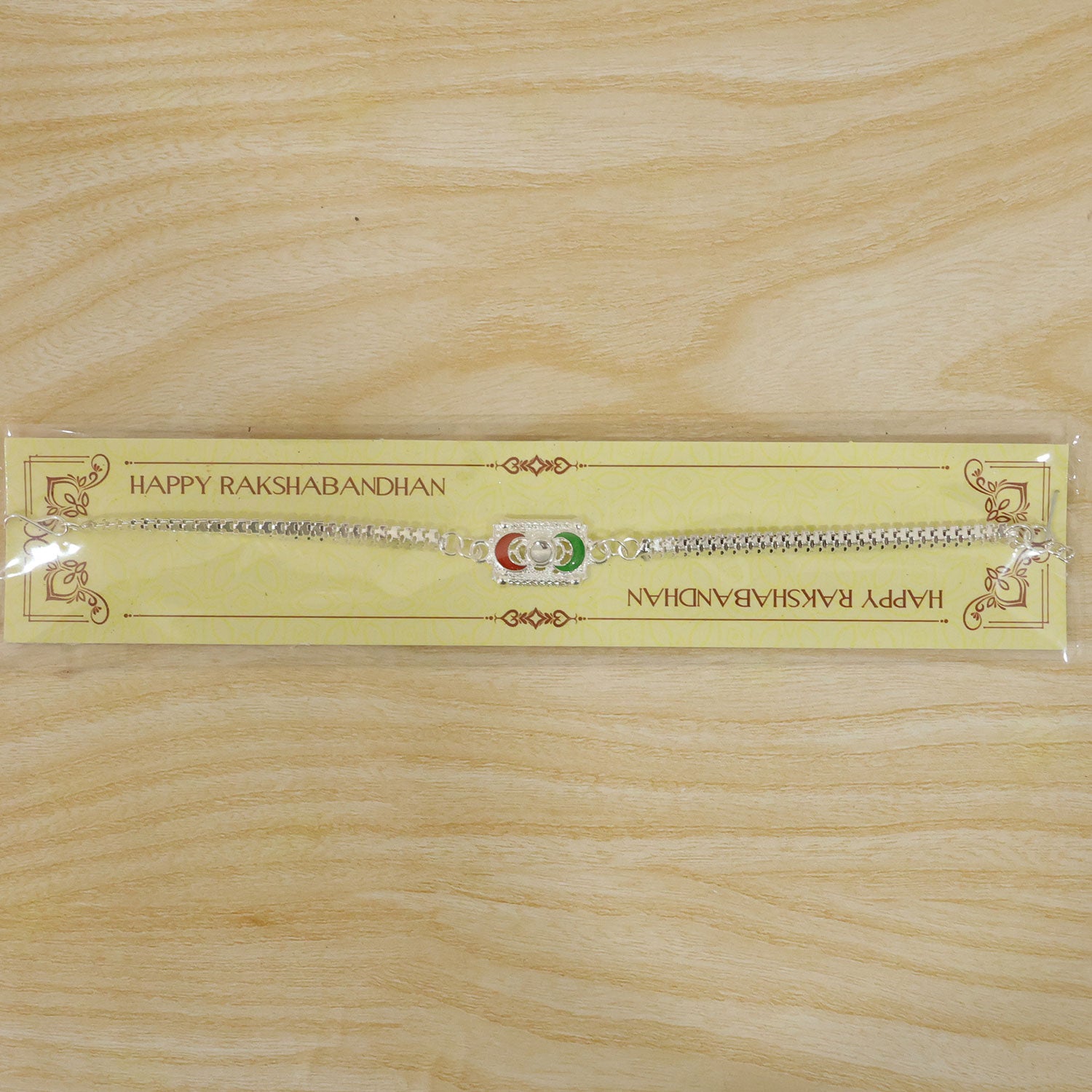 A silver-colored bracelet with a rectangular centerpiece featuring red and green crescent moon shapes, packaged on a yellow