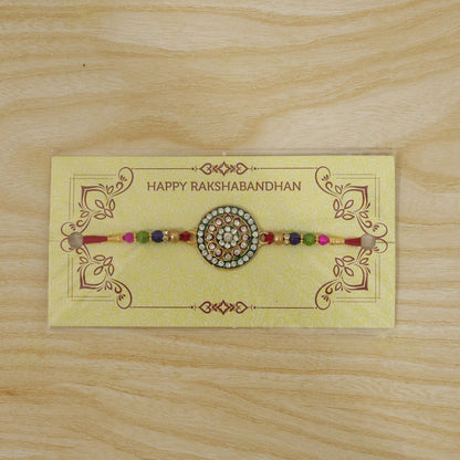 A red thread bracelet with a gold, green, and red circular centerpiece adorned with clear stones, displayed on a yellow card