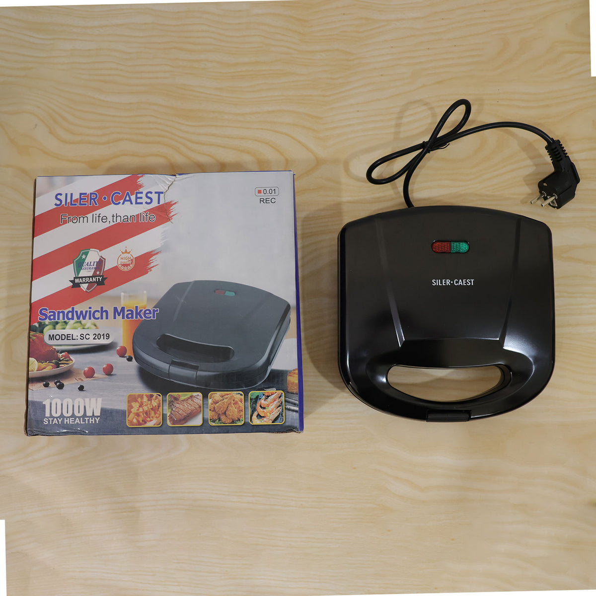 A black sandwich maker with its power cord and a product box, both resting on a light wooden surface.