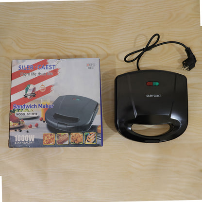 A black sandwich maker with its power cord and a product box, both resting on a light wooden surface.