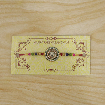 A gold and green circular Rakhi with clear stones and colorful beads is presented on a yellow
