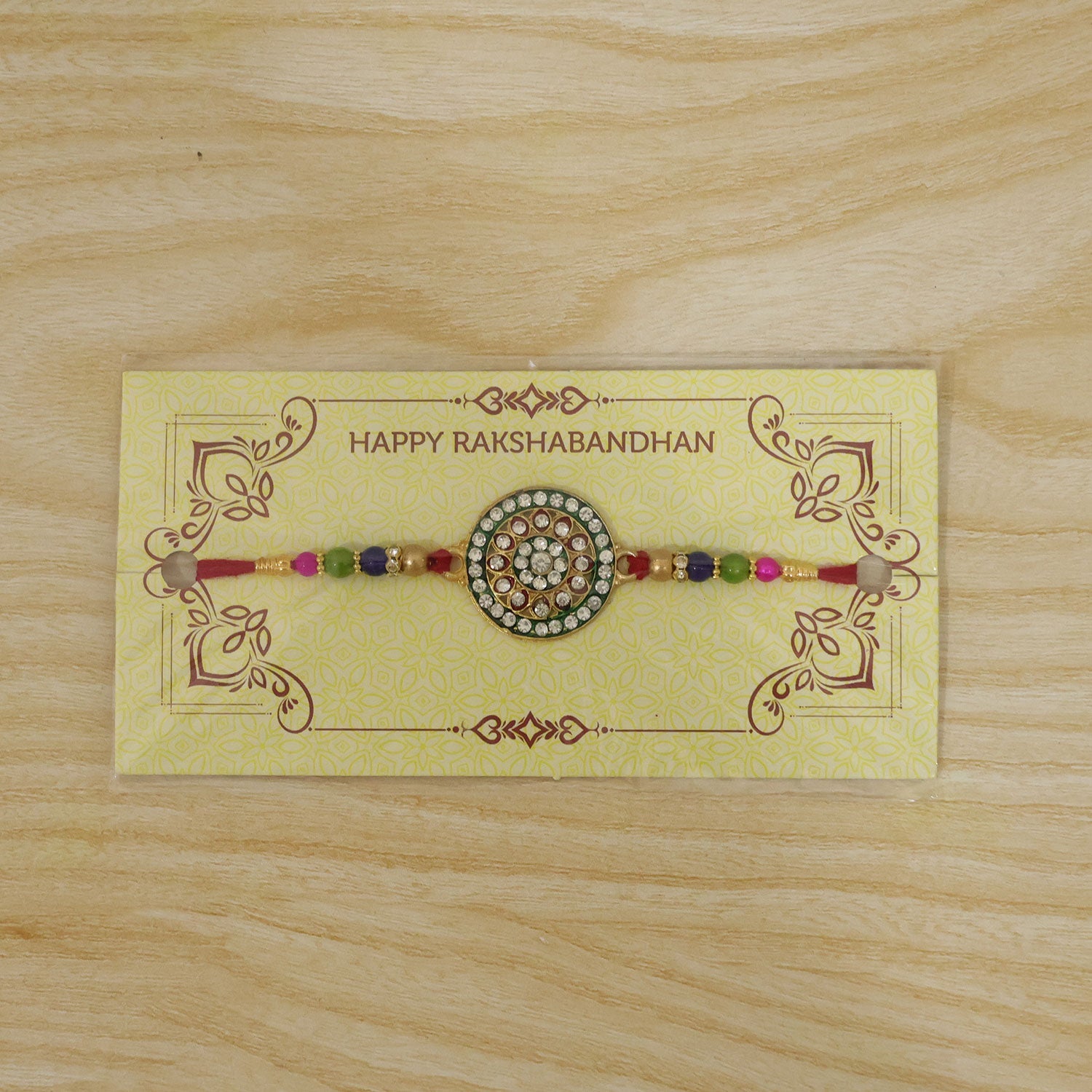 A red thread bracelet with a gold-toned kundan centerpiece and colorful beads is displayed on a yellow card with