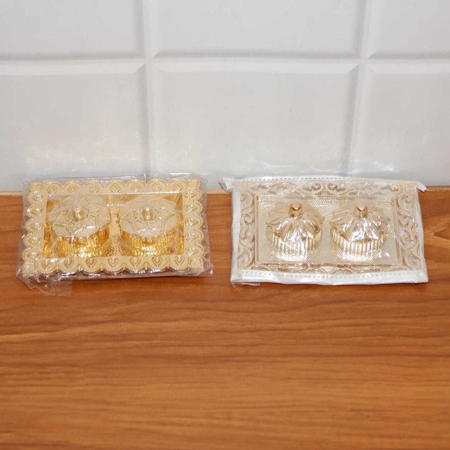 Two ornate rectangular pooja thalis, one gold and one silver, both in clear plastic packaging, on a wooden surface.