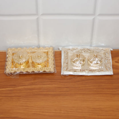 Two ornate rectangular pooja thalis, one gold and one silver, both in clear plastic packaging, on a wooden surface.