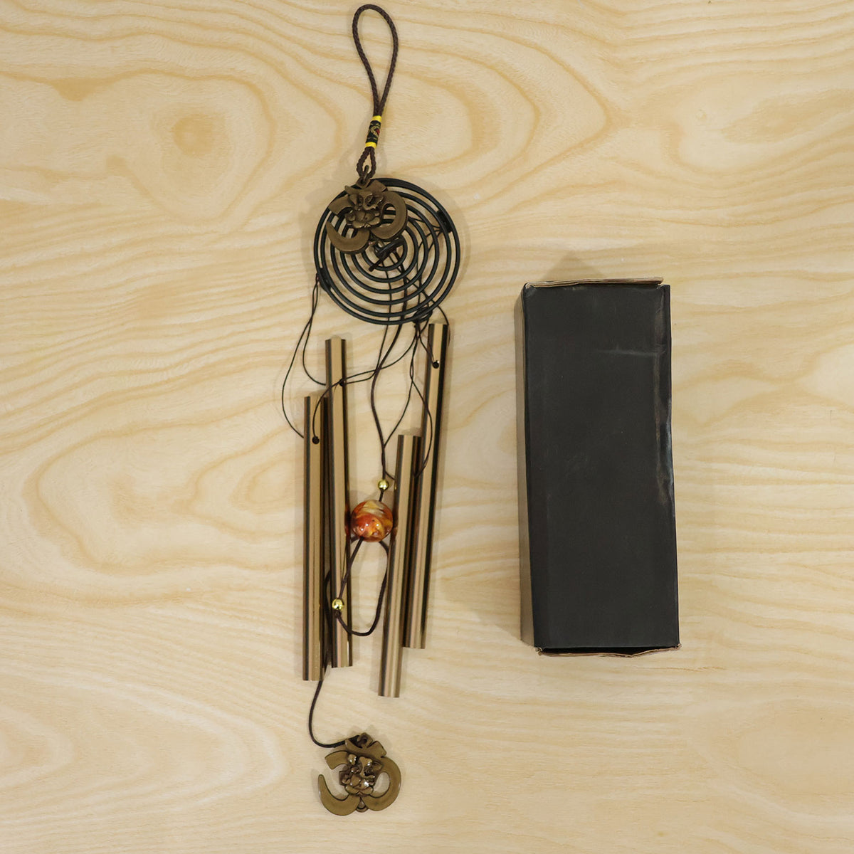 A metal wind chime with five gold-toned tubes and Om symbols, laid flat on a wooden surface next to a black protective box.