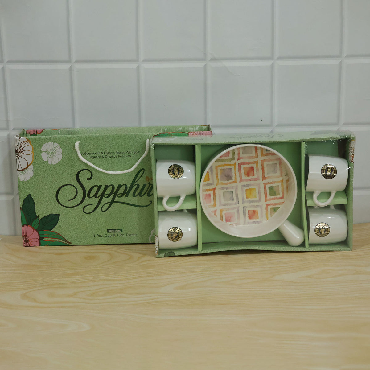 A green gift box containing a five-piece ceramic set, with four white mugs and a colorful patterned platter, sits on a