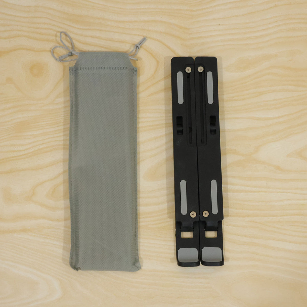 A folded black laptop stand with grey pads lies beside a light grey drawstring carrying bag on a wooden surface.