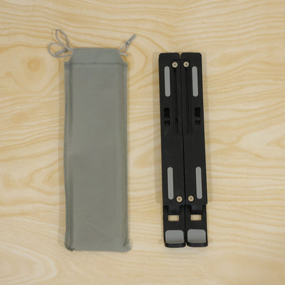 A folded black laptop stand with grey pads lies beside a light grey drawstring carrying bag on a wooden surface.