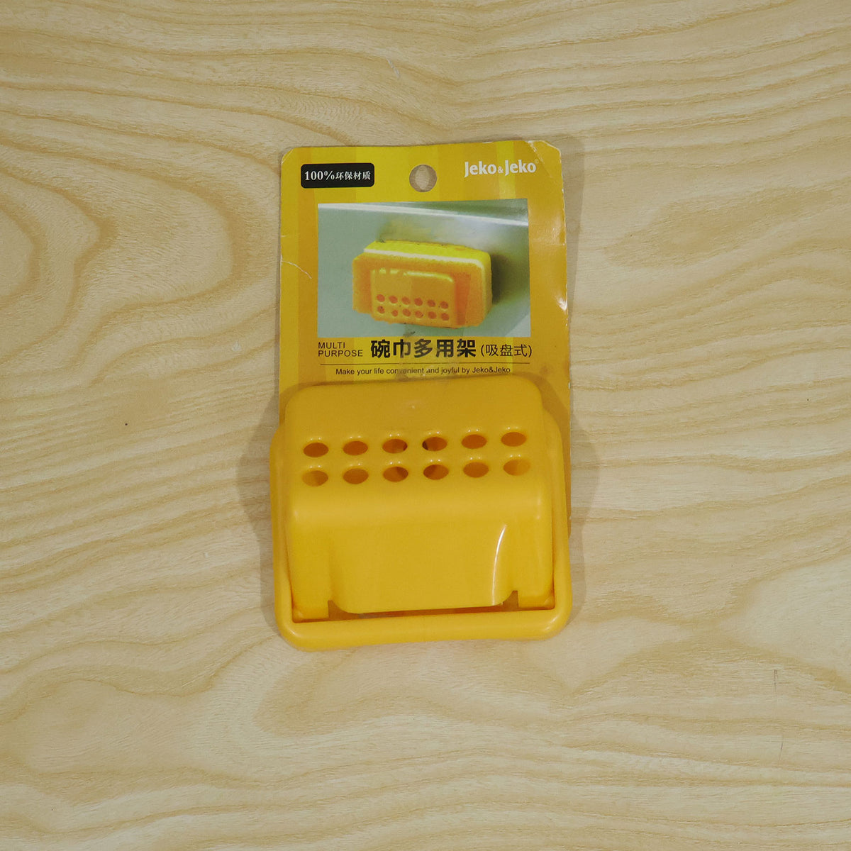 A yellow plastic sponge holder in its original packaging, lying on a light wooden surface.