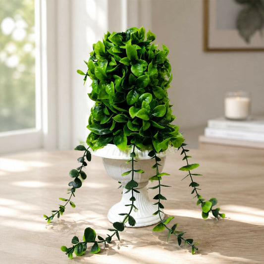 Vibrant green artificial plant in a decorative white urn-shaped pot on a light wooden table near a window.