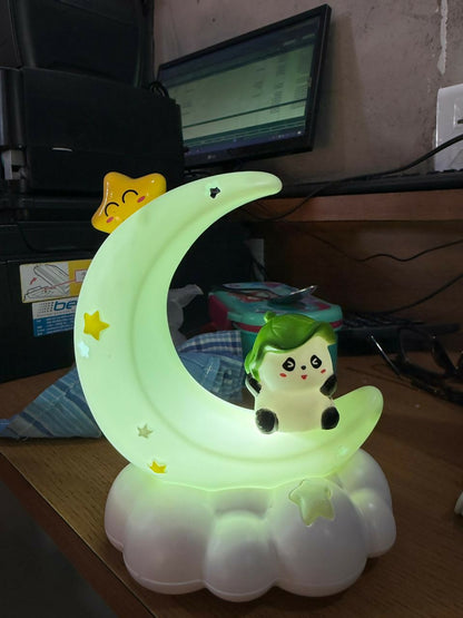 A green crescent moon night light with a character and star, illuminating a cluttered desk with a computer monitor in the