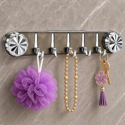 A gray transparent wall rack with five silver folding hooks holds a purple loofah, a gold beaded necklace, and a key with a