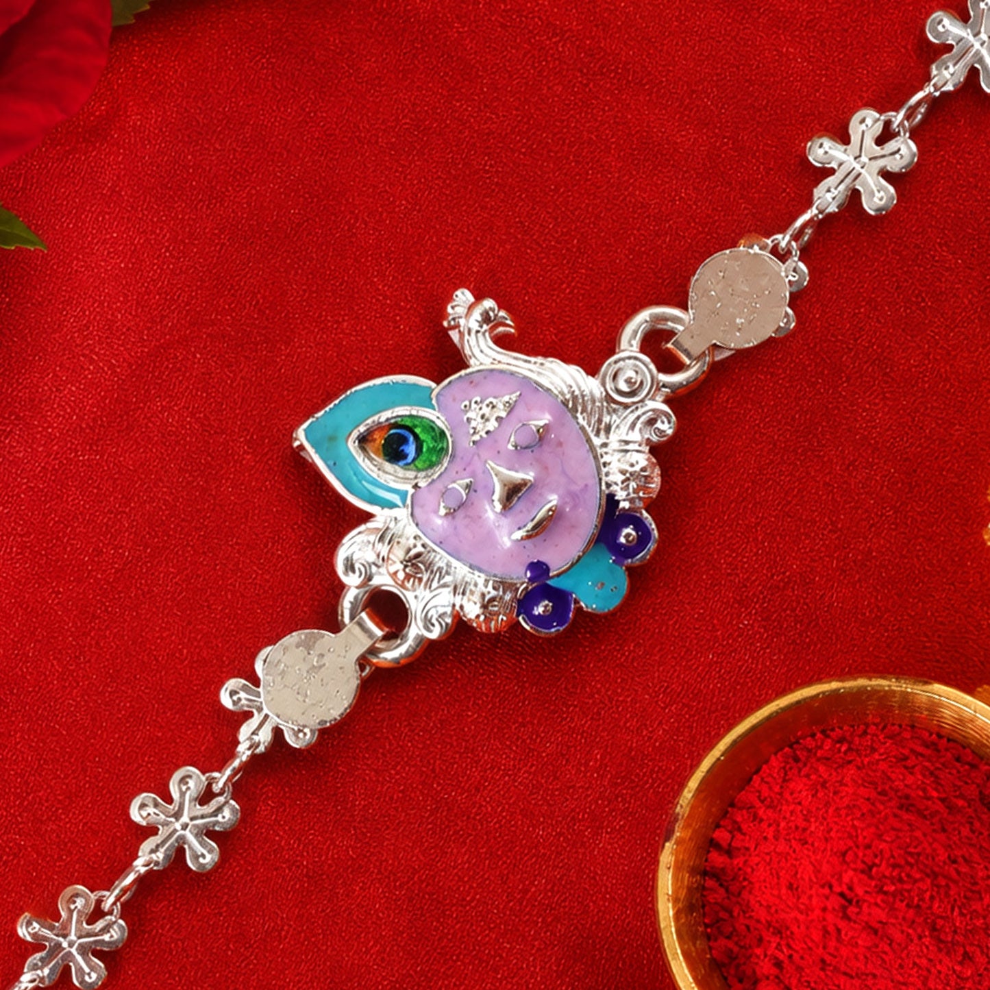 Silver rakhi with a purple enamel Krishna face and peacock feather charm on a vibrant red background with kumkum.