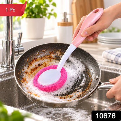 A hand holds a pink and white dish brush, scrubbing a soiled frying pan with soapy water in a stainless steel sink.