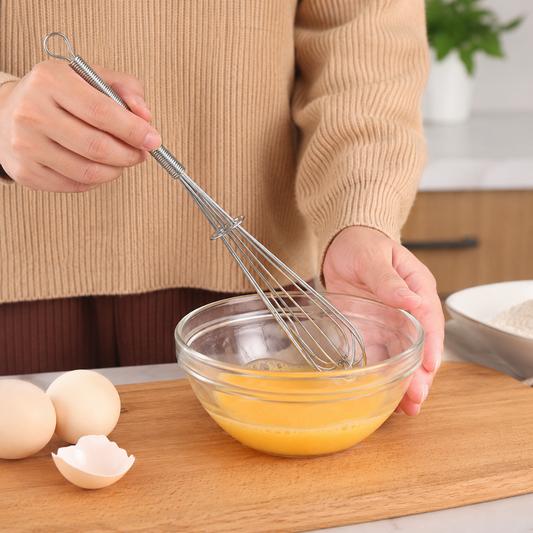 A person in a beige sweater uses a small silver whisk to beat eggs in a clear glass bowl on a wooden board.