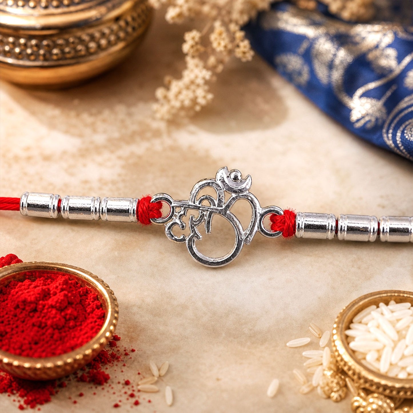 A silver Om symbol with Ganesha's face is centered on a red thread, flanked by silver beads, with red powder and rice