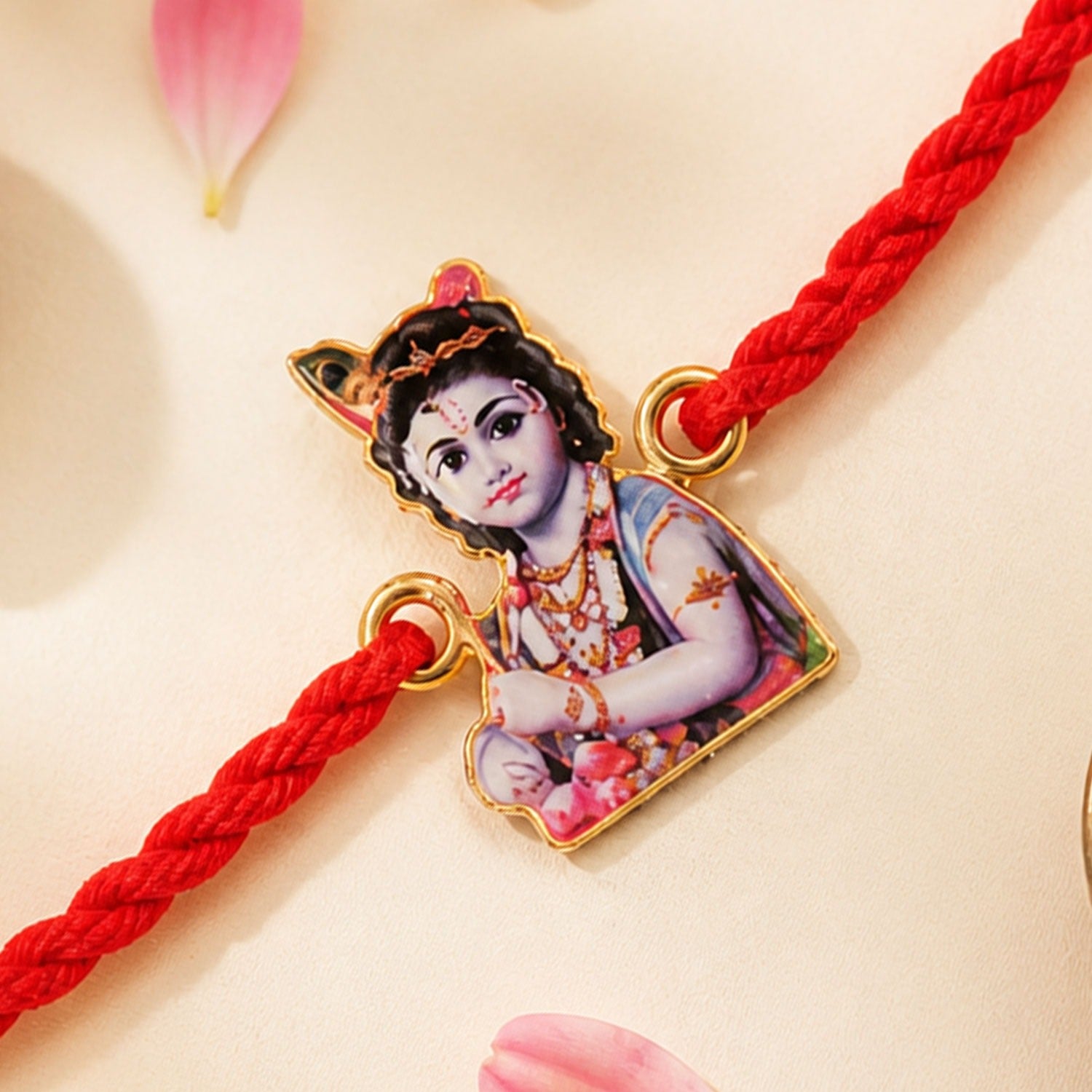 Close-up of a gold-framed charm with baby Krishna on a braided red thread, with pink petals nearby.